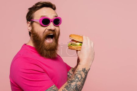 thrilled man in trendy sunglasses opening mouth near tasty burger isolated on pink