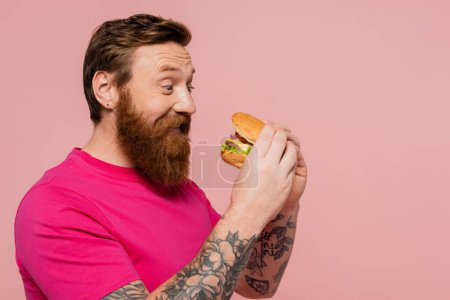Foto de Hombre barbudo complacido en camiseta magenta sosteniendo sabrosa hamburguesa aislada en rosa - Imagen libre de derechos