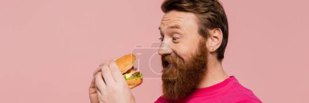 Foto de Hombre barbudo emocionado mirando deliciosa hamburguesa aislada en rosa, bandera - Imagen libre de derechos