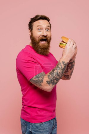 excited tattooed man in bright t-shirt looking at camera while holding hamburger isolated on pink