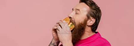 Foto de Hombre barbudo con los ojos cerrados comiendo deliciosa hamburguesa aislada en rosa, pancarta - Imagen libre de derechos