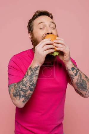 hombre tatuado complacido con los ojos cerrados comiendo deliciosa hamburguesa aislada en rosa