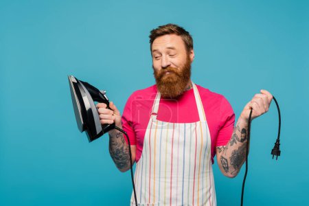 skeptical bearded man in striped apron looking at iron and smiling isolated on blue