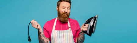 Photo for Smiling and skeptical man in striped apron holding electric iron isolated on blue, banner - Royalty Free Image
