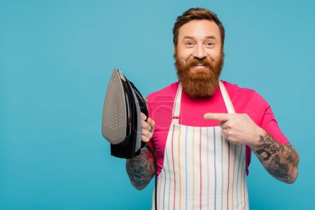 happy tattooed man in striped apron looking at camera while pointing at iron isolated on blue