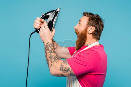 Téléchargez les photos : Vue latérale de l'homme tatoué barbu dans le tablier tenant fer et hurlant isolé sur bleu - en image libre de droit