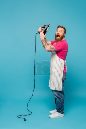 full length of thrilled man with iron shouting and looking at camera on blue background
