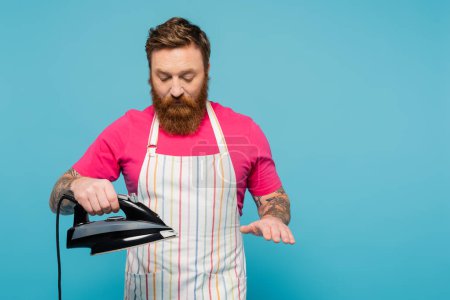 bearded man in magenta t-shirt and striped apron holding iron isolated on blue
