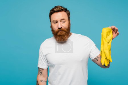 Foto de Hombre barbudo disgustado en camiseta blanca con guantes de goma amarillos aislados en azul - Imagen libre de derechos