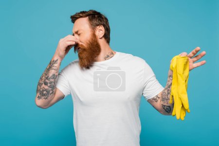 angeekelter bärtiger Mann im weißen T-Shirt, der die Nase stopft, während er stinkende Gummihandschuhe isoliert auf blauem Grund hält