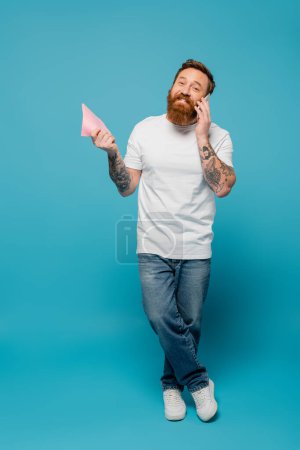 longitud completa de hombre barbudo feliz en camiseta blanca y pantalones vaqueros sosteniendo trapo y hablando en el teléfono celular sobre fondo azul