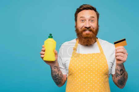 Foto de Cheerful tattooed man in yellow apron showing detergent and credit card isolated on blue - Imagen libre de derechos