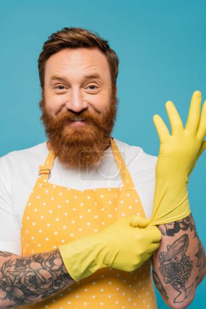 Téléchargez les photos : Joyeux homme barbu en tablier à pois portant des gants en caoutchouc jaune et regardant la caméra isolée sur bleu - en image libre de droit
