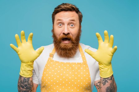 excited bearded man in apron and yellow rubber gloves showing scaring gesture isolated on blue