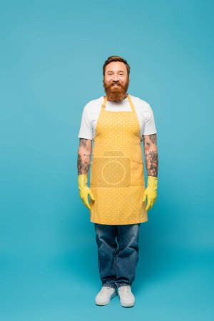 full length of bearded tattooed man in yellow apron and rubber gloves smiling at camera on blue background