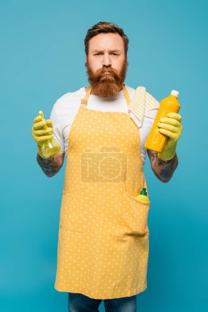 displeased man in yellow apron puffing cheeks while standing with detergent and spray bottle isolated on blue