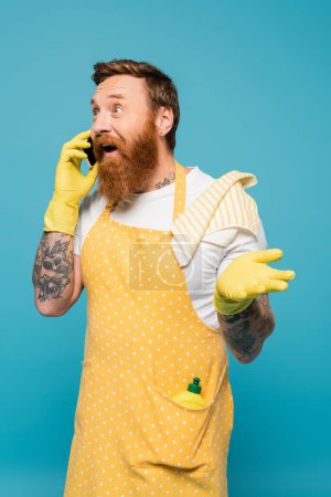 Téléchargez les photos : Excité homme barbu à pois tablier parlant sur smartphone et pointant avec la main isolé sur bleu - en image libre de droit