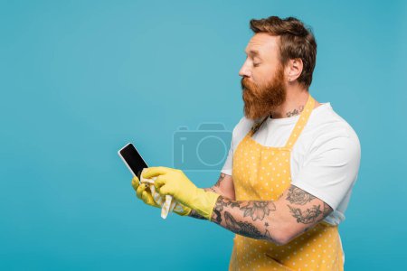 Foto de Bearded man in white t-shirt and yellow apron wiping smartphone with blank screen isolated on blue - Imagen libre de derechos