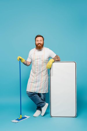fröhlich tätowierter Mann in gelben Gummihandschuhen und gestreifter Schürze, der einen Wischmopp neben einer riesigen Smartphone-Vorlage auf blauem Hintergrund hält