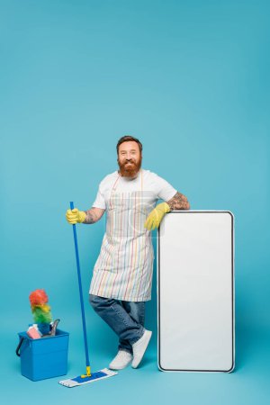 hombre feliz en delantal rayado y guantes de goma amarilla de pie con fregona cerca de la plantilla de teléfono enorme y suministros de limpieza sobre fondo azul