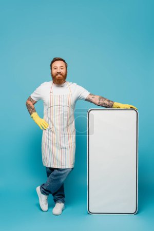 heureux homme barbu en tablier rayé et gants en caoutchouc jaune posant avec la main sur la hanche près de gabarit de téléphone énorme sur fond bleu