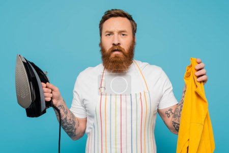 Photo for Tense bearded man in striped apron holding yellow cloth and iron isolated on blue - Royalty Free Image