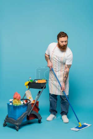Téléchargez les photos : Homme barbu sérieux sur toute la longueur avec sol de nettoyage de serpillière près du chariot avec des fournitures de nettoyage sur fond bleu - en image libre de droit