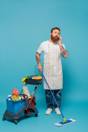 serious man with mop talking on mobile phone near cart with cleaning supplies on blue background