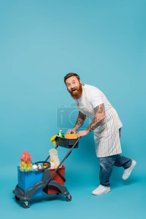 full length of excited tattooed man looking at camera near cart with cleaning supplies on blue background