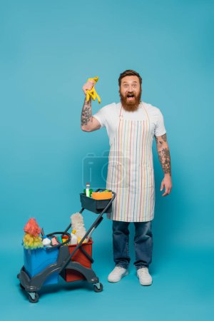 amazed bearded man in apron holding rubber gloves near cart with cleaning supplies on blue background