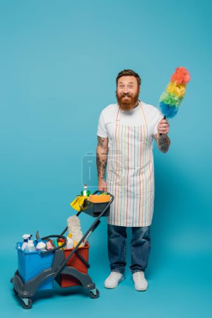 Téléchargez les photos : Homme gai dans tablier posant avec brosse à poussière près du chariot avec des fournitures de nettoyage sur fond bleu - en image libre de droit