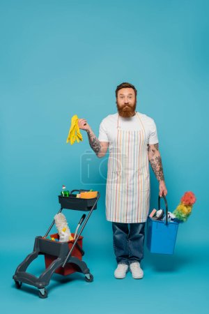 positiver bärtiger Mann mit Gummihandschuhen und Eimer, der in die Kamera neben dem Einkaufswagen mit Putzmitteln auf blauem Hintergrund blickt