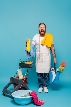 hombre barbudo estresado en delantal rayado gritando cerca de lavandería y artículos de limpieza sobre fondo azul