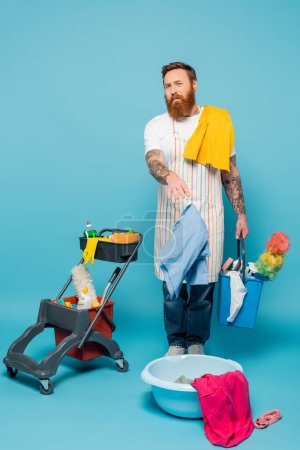 Unzufriedener Mann mit Kleidungsstücken und Eimer zeigt auf Wäschekorb neben Einkaufswagen mit Putzmitteln auf blauem Hintergrund
