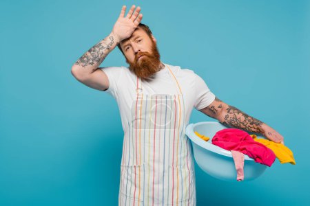 exhausted bearded man holding hand near forehead while standing with laundry bowl isolated on blue