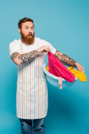 discouraged bearded man in apron holding laundry bowl and looking at camera isolated on blue
