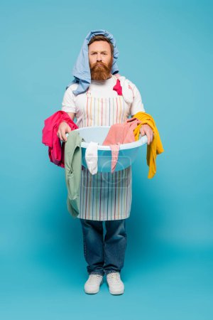 Unzufriedener bärtiger Mann mit Kleidungsstücken auf dem Kopf hält Wäscheschale in der Hand und blickt in die Kamera auf blauem Hintergrund