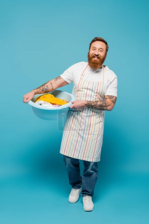 Photo for Full length of happy tattooed man in striped apron holding laundry bowl and smiling at camera on blue background - Royalty Free Image