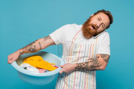 Photo for Exhausted tattooed man talking on mobile phone while holding laundry bowl isolated on blue - Royalty Free Image