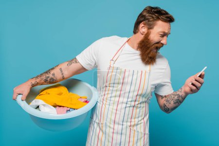 shocked man in apron screaming while holding laundry bowl and looking at smartphone isolated on blue