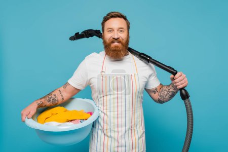 Photo for Cheerful bearded man with laundry bowl and vacuum cleaner looking at camera isolated on blue - Royalty Free Image