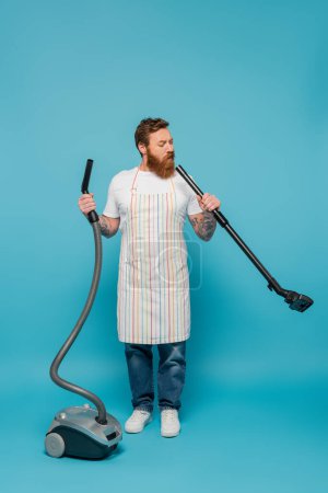full length of thoughtful man in striped apron looking in tube of vacuum cleaner on blue background
