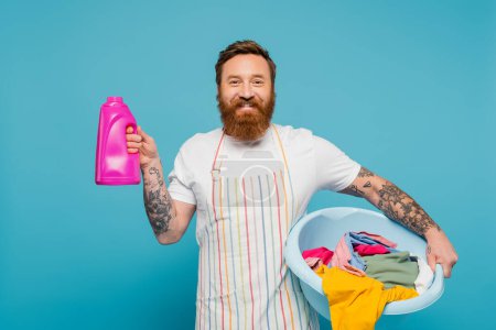 positive tattooed man with washing gel and laundry bowl looking at camera isolated on blue