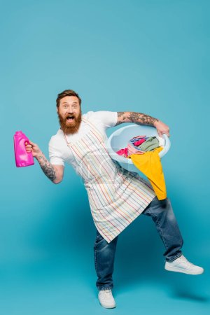 full length of excited bearded man posing with washing gel and laundry bowl on blue background