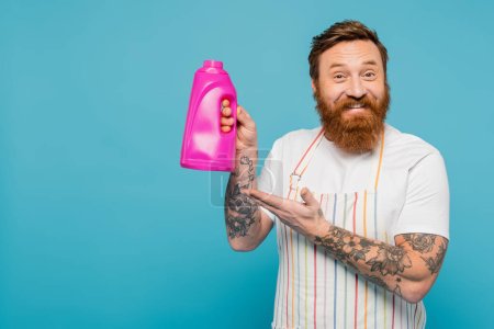 Téléchargez les photos : Joyful bearded man in striped apron pointing at bottle with washing gel isolated on blue - en image libre de droit