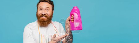 Téléchargez les photos : Happy bearded man in apron pointing at bottle with washing gel isolated on blue, banner - en image libre de droit