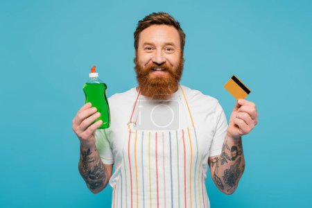Téléchargez les photos : Happy bearded man with dishwashing gel and credit card smiling at camera isolated on blue - en image libre de droit