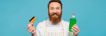 Téléchargez les photos : Happy bearded man in white t-shirt and apron holding credit card and dishwashing gel isolated on blue, banner - en image libre de droit