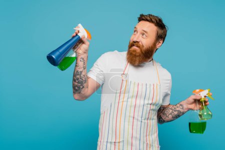 Foto de Hombre barbudo alegre con botellas de spray sonriendo y mirando hacia otro lado aislado en azul - Imagen libre de derechos