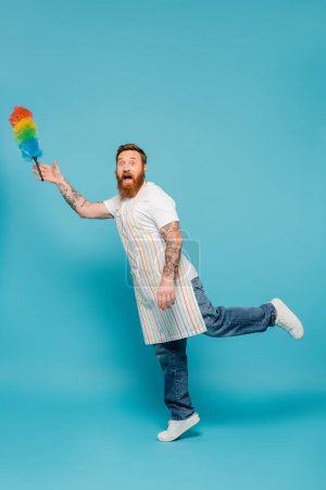 full length of amazed bearded man in striped apron looking at camera near levitating dust brush on blue background